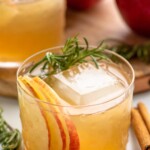 Apple Cider Cocktail served in a glass with ice, garnished with rosemary sprig and apple slices. Cinnamon sticks, rosemary, and apples beside. - 6