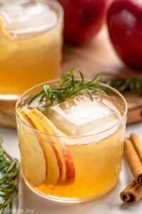 Apple Cider Cocktail served in a glass with ice, garnished with rosemary sprig and apple slices. Cinnamon sticks, rosemary, and apples beside. - 5