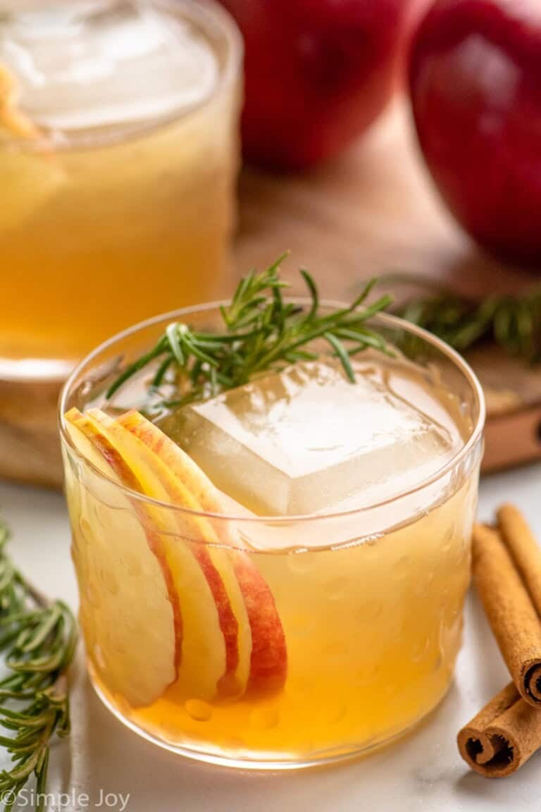 Apple Cider Cocktail served in a glass with ice, garnished with rosemary sprig and apple slices. Cinnamon sticks, rosemary, and apples beside.