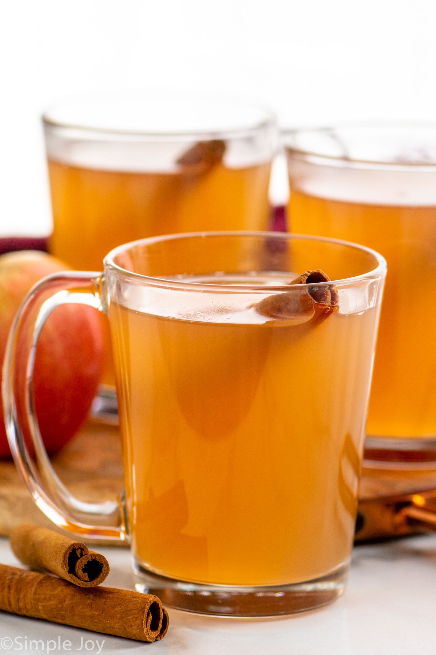Side view of a mugs of Apple Cider recipe garnished with cinnamon sticks, with cinnamon sticks and apples beside.