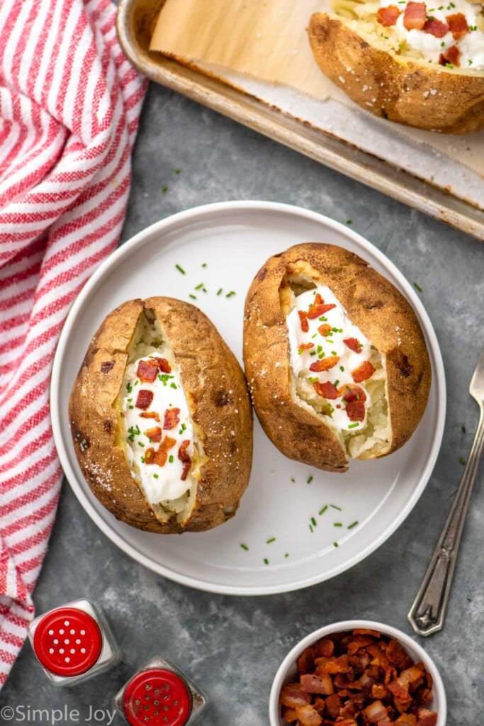 Overhead view of a plate with baked potatoes garnished with sour cream and bacon bits. Salt, pepper, and bacon pieces beside. - 5