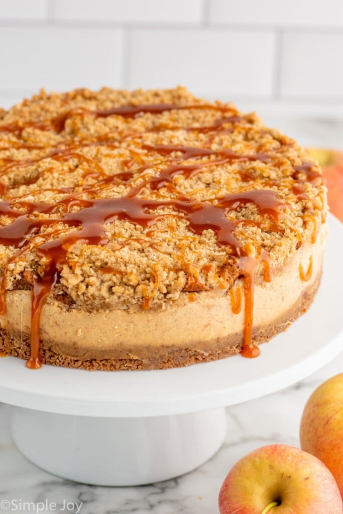 Caramel Apple Cheesecake garnished with caramel drizzle served on a cake platter with apples beside. - 15