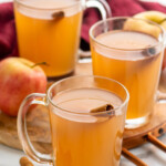 Mugs of Apple Cider garnished with cinnamon sticks, cinnamon sticks and apples beside. - 10