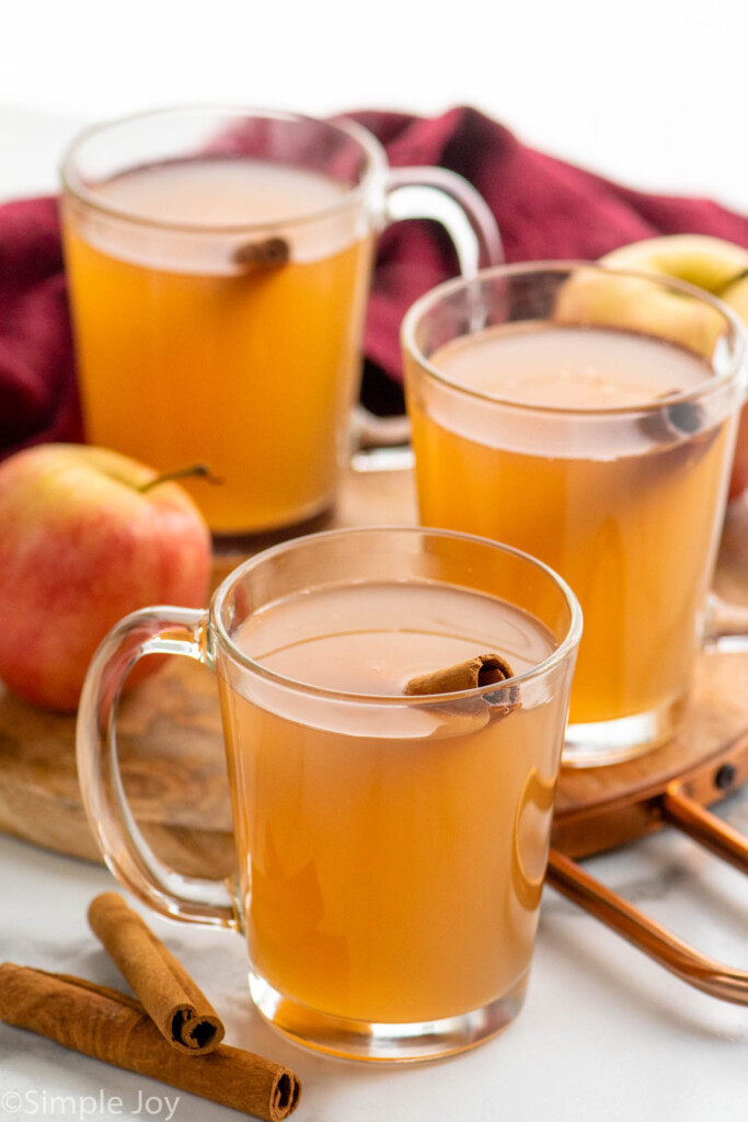 Mugs of Apple Cider garnished with cinnamon sticks, cinnamon sticks and apples beside. - 8