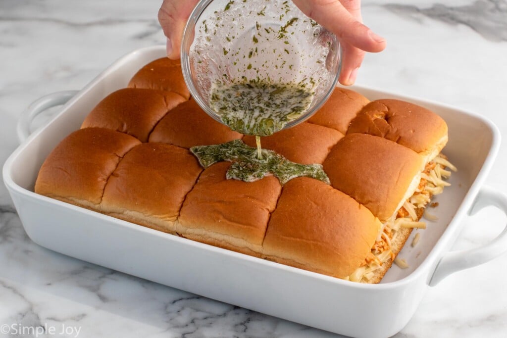 Photo of a baking dish of Buffalo Chicken Sliders in a baking dish with person's hand pouring seasonings over top of rolls.