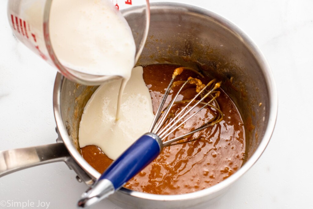 Measuring cup of cream being added to pot of caramel sauce recipe with whisk for mixing
