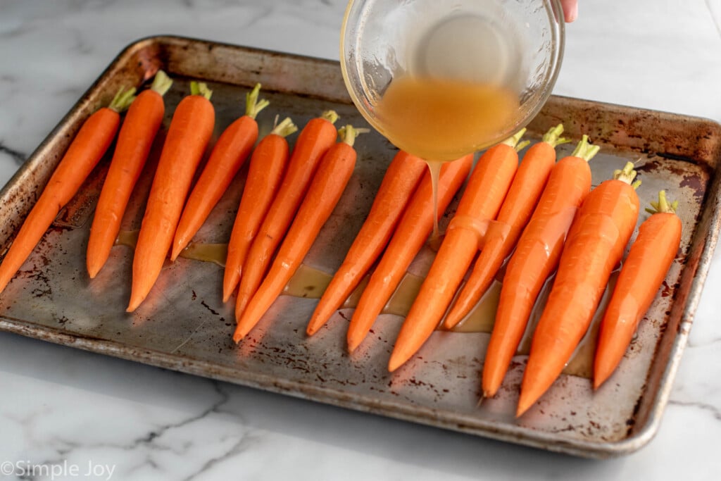 Photo of bowl of glaze being poured over whole carrots on a baking sheet for Honey Roasted Carrots recipe