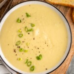 Overhead view of a bowl of potato leek soup garnished with chopped green onions, bread and spoons beside. - 11