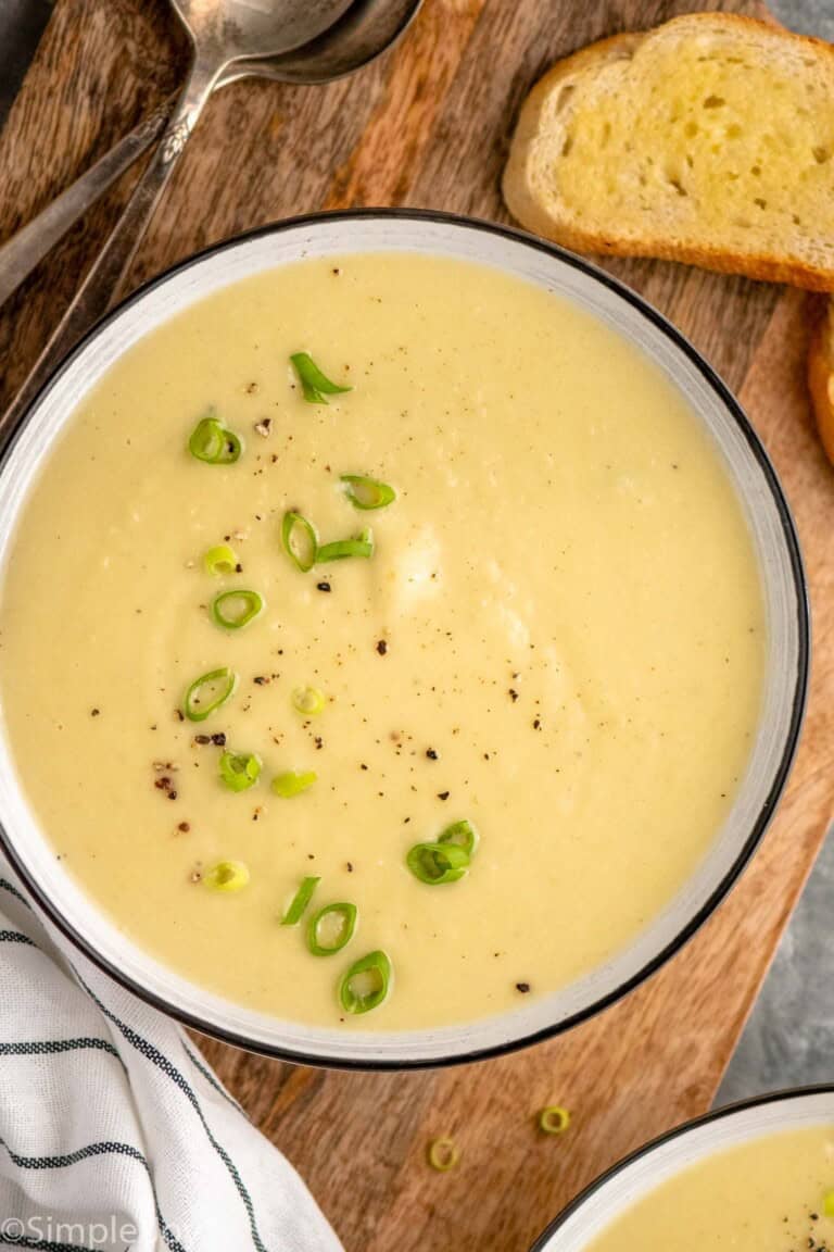 Overhead view of a bowl of potato leek soup garnished with chopped green onions, bread and spoons beside.