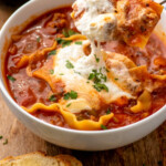 Pinterest graphic for Lasagna Soup recipe. Image shows a bowl of Lasagna Soup with a spoon lifting a bite. Glass of red wine and bread beside. Text says, "the best Lasagna Soup simplejoy.com."