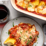Pinterest graphic for manicotti recipe Text says, "delicious manicotti recipe simplejoy.com." Image shows overhead view of a plate of manicotti garnished with parsley flakes, glass of red wine beside.