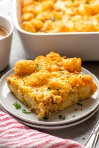 Piece of tater tot breakfast casserole served on a plate with baking dish of tater tot breakfast casserole beside.