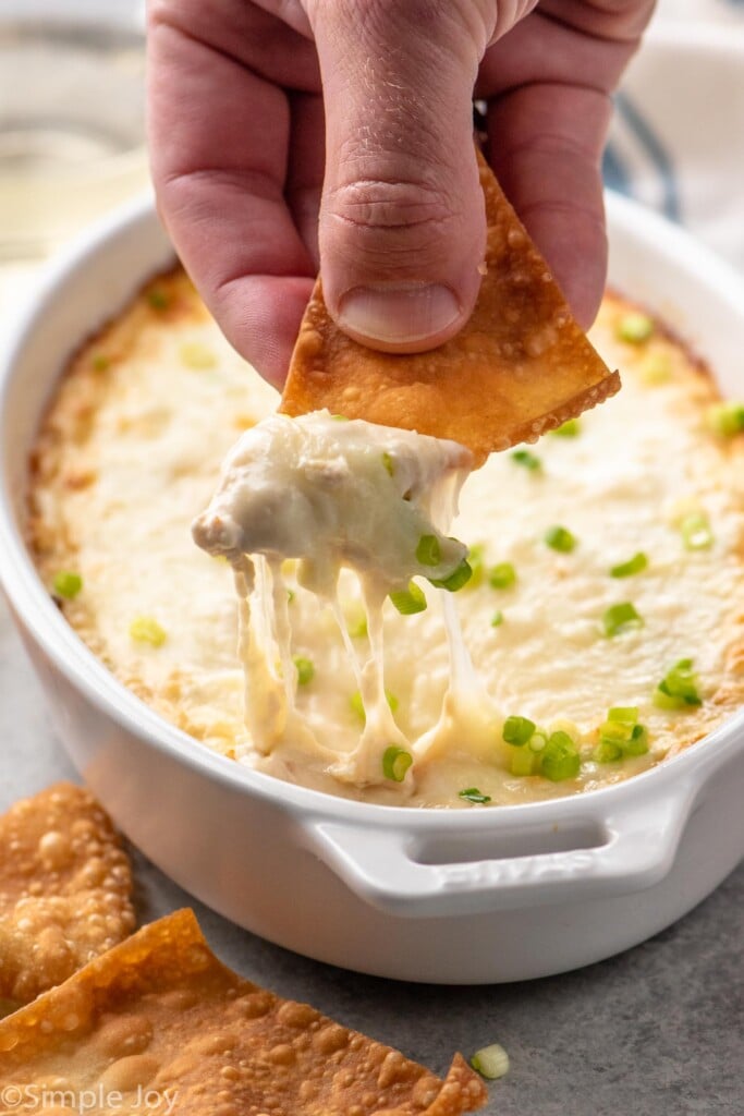 Person's hand dipping pita chip into baking dish of crab rangoon dip, more pita chips beside/ - 8