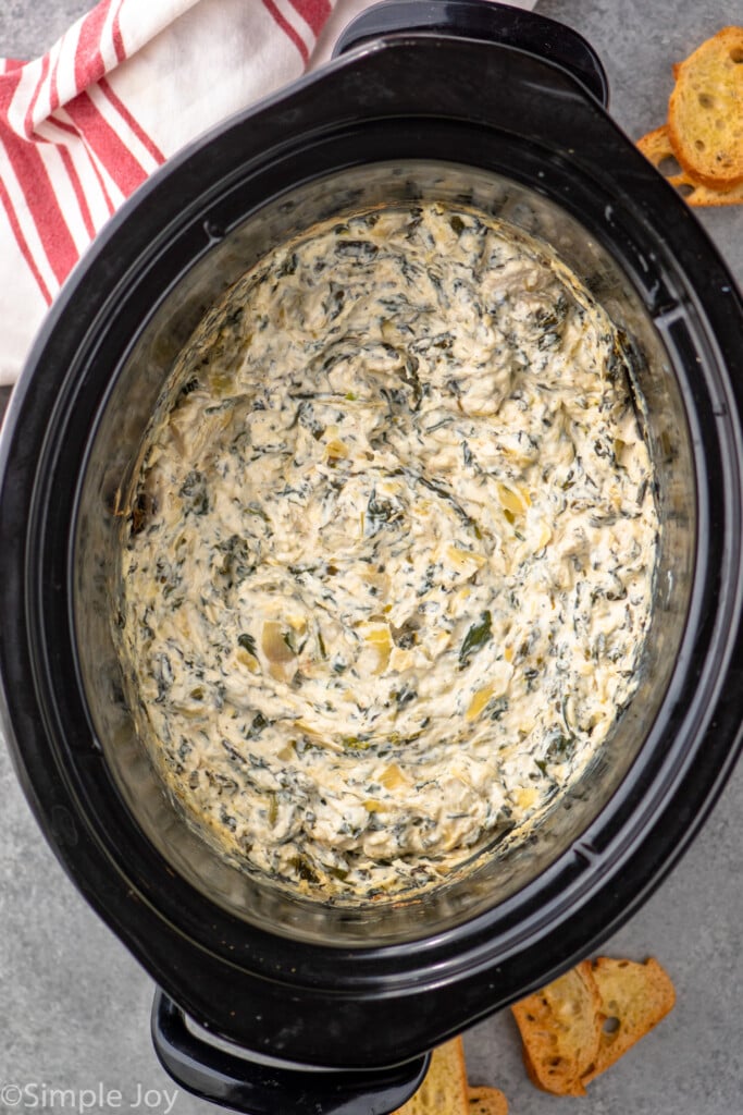 Overhead view of a crock pot of Crockpot Spinach Artichoke Dip with slices of toasted bread beside. - 7