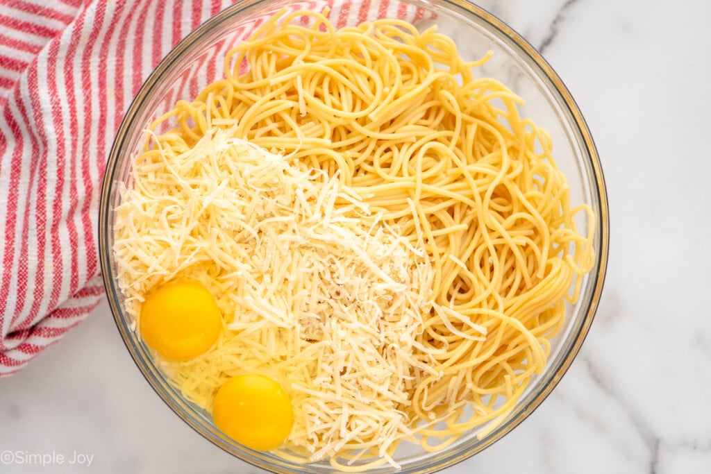 Overhead view of a glass mixing bowl of spaghetti noodles, cheese, and eggs for Baked Spaghetti recipe.