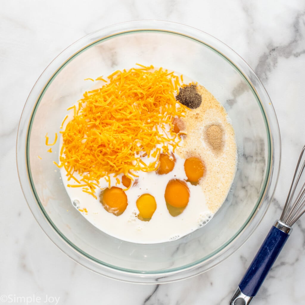 Overhead view of a glass mixing bowl of ingredients for tater tot breakfast casserole recipe, with whisk beside.