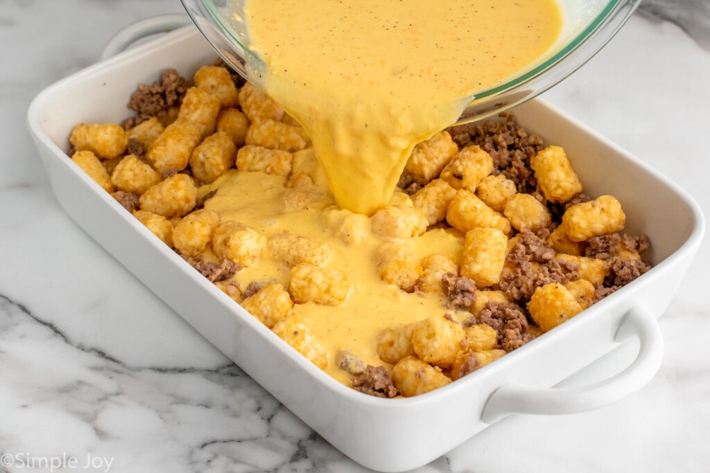 Bowl of tater tot breakfast casserole sauce being poured into baking dish of tater tots and ground sausage for tater tot breakfast casserole recipe.