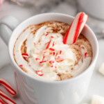 Mug of Peppermint Hot Chocolate garnished with whipped cream and candy canes, marshmallows and candy canes beside. - 8