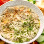 View of a bowl of Baba Ganoush garnished with parsley, surrounded be pita chips and sliced vegetables for dipping. - 11
