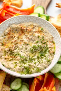 View of a bowl of Baba Ganoush garnished with parsley, surrounded be pita chips and sliced vegetables for dipping. - 10