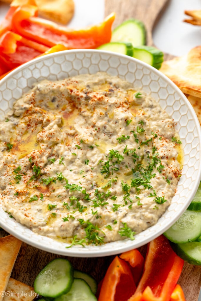 View of a bowl of Baba Ganoush garnished with parsley, surrounded be pita chips and sliced vegetables for dipping. - 3