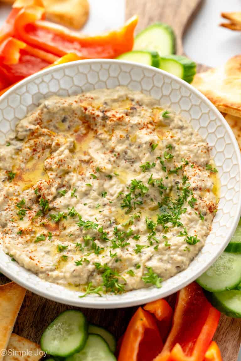 View of a bowl of Baba Ganoush garnished with parsley, surrounded be pita chips and sliced vegetables for dipping.