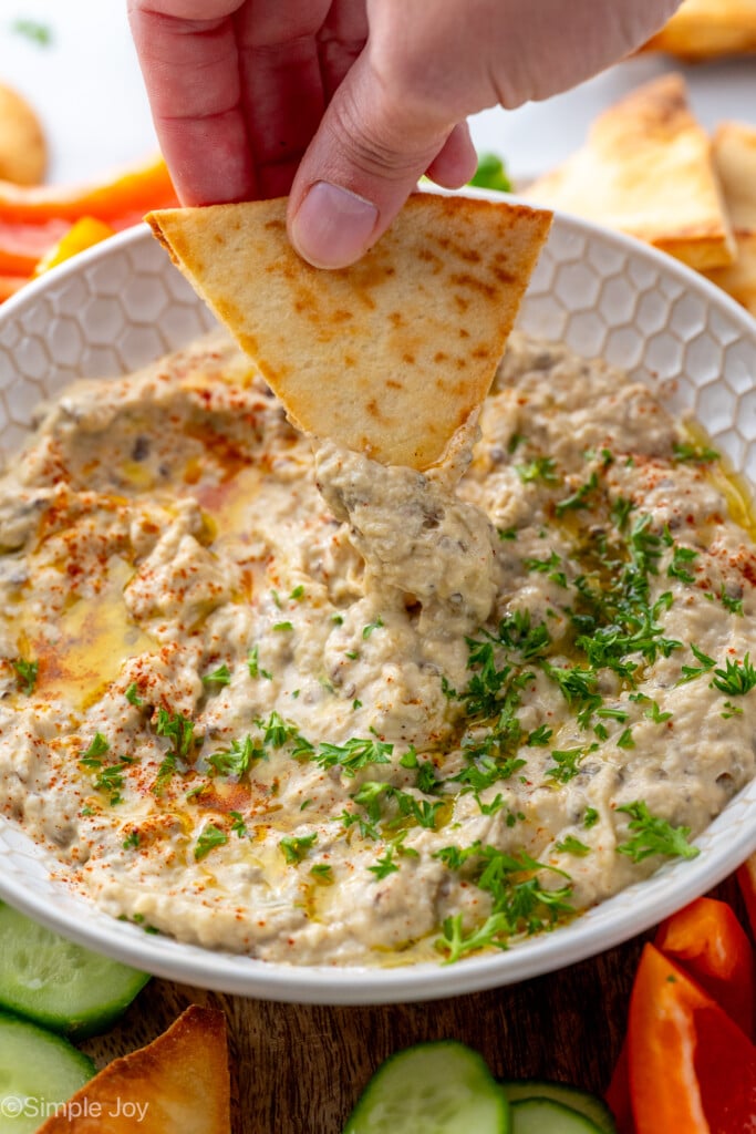 Person's hand dipping pita chip into bowl of Baba Ganoush garnished with parsley. - 9
