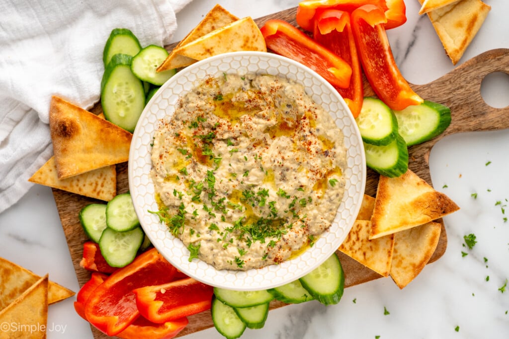 Overhead view of a bowl of Baba Ganoush garnished with parsley, surrounded be pita chips and sliced vegetables for dipping. - 8