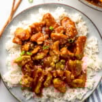 Overhead view of a plate of rice and Bourbon Chicken garnished with chopped green onions and sesame seeds, chopsticks beside.