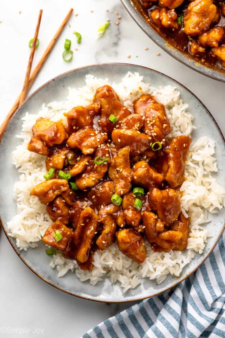 Overhead view of a plate of rice and Bourbon Chicken garnished with chopped green onions and sesame seeds, chopsticks beside.