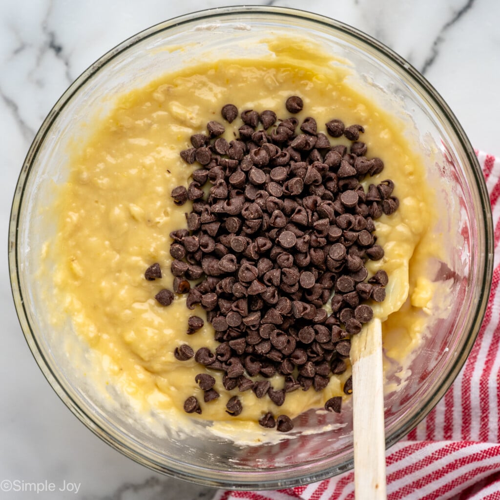 Overhead view of a glass mixing bowl of ingredients for banana chocolate chip muffins recipe with a spatula for stirring. - 10