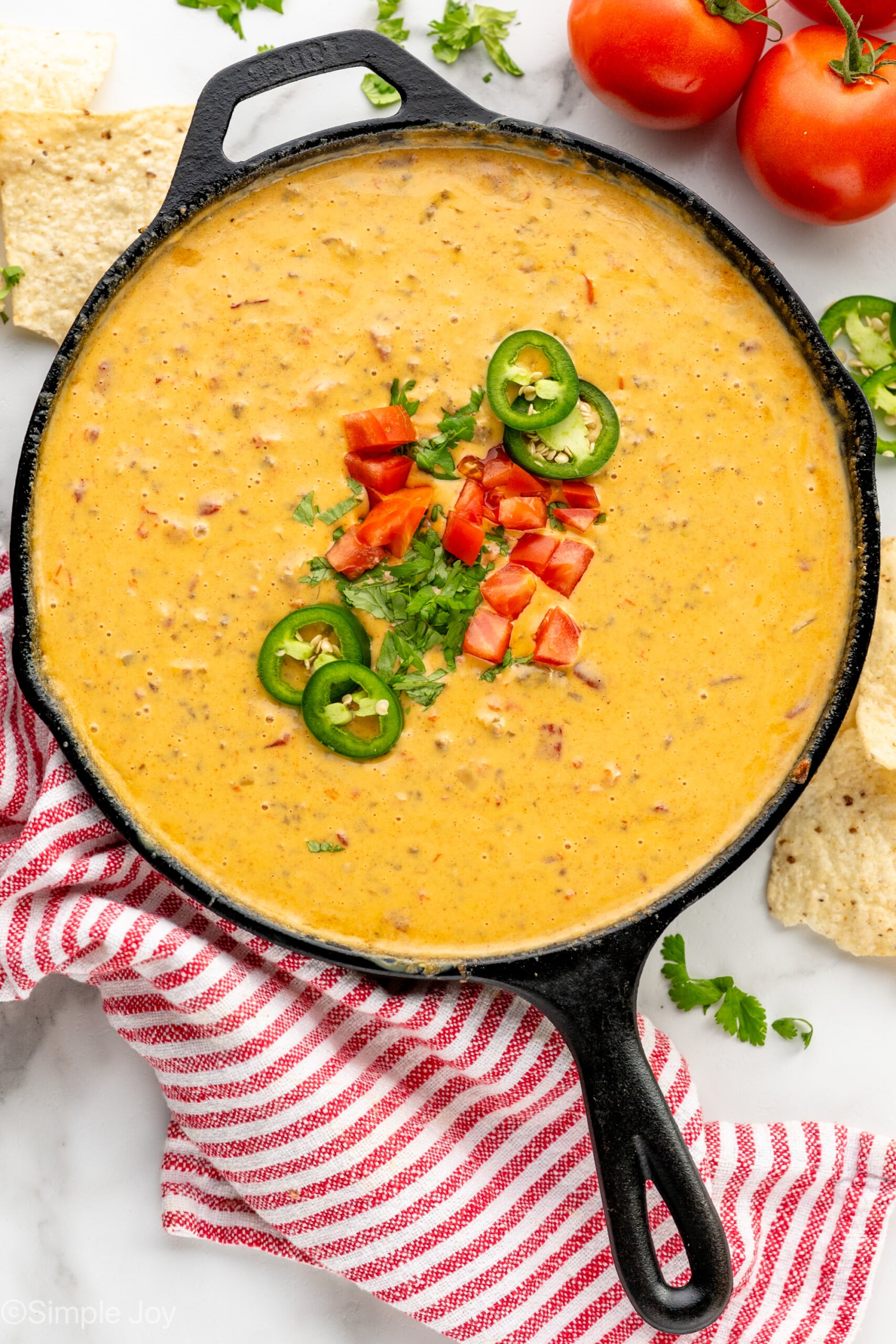 Overhead view of a skillet of Rotel Dip garnished with cilantro, chopped tomatoes, and jalapenos. Tomatoes, jalapenos, and tortilla chips beside