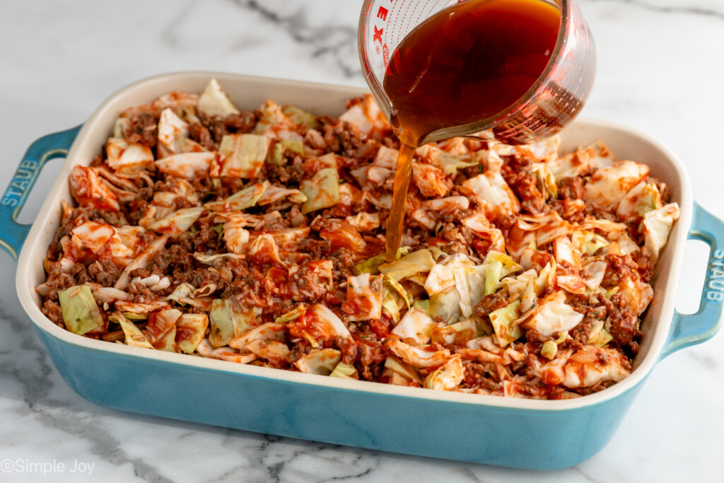 Glass measuring cup of beef broth being poured over baking dish of ingredients for cabbage roll casserole recipe