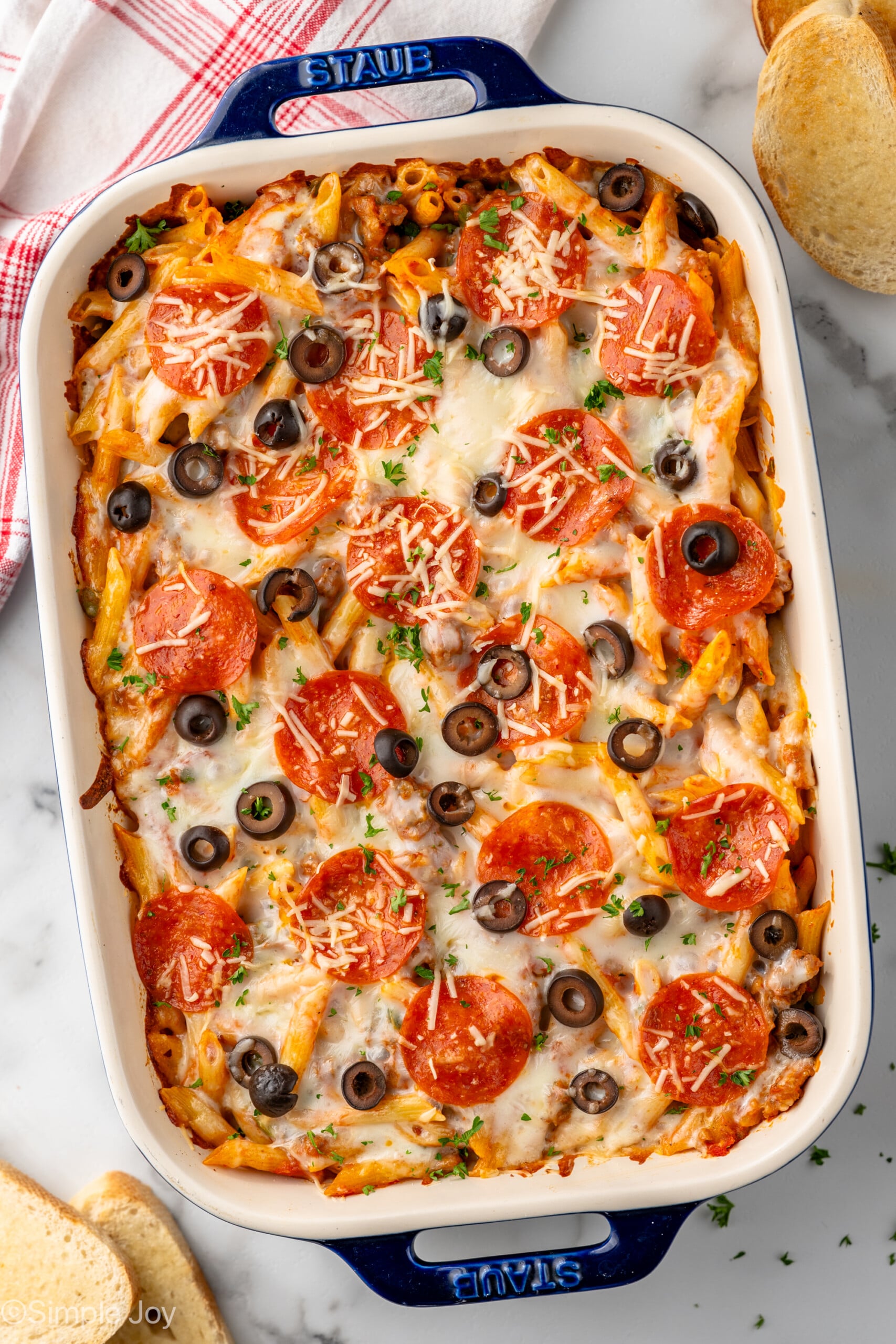 Overhead view of baking dish of Pizza Casserole garnished with shredded cheese, pepperoni, and black olives. Toast beside.
