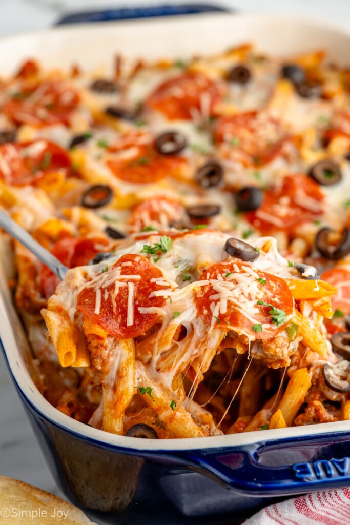 Side view of spoon serving Pizza Casserole from baking dish. Pizza Casserole is topped with black olives and pepperoni.