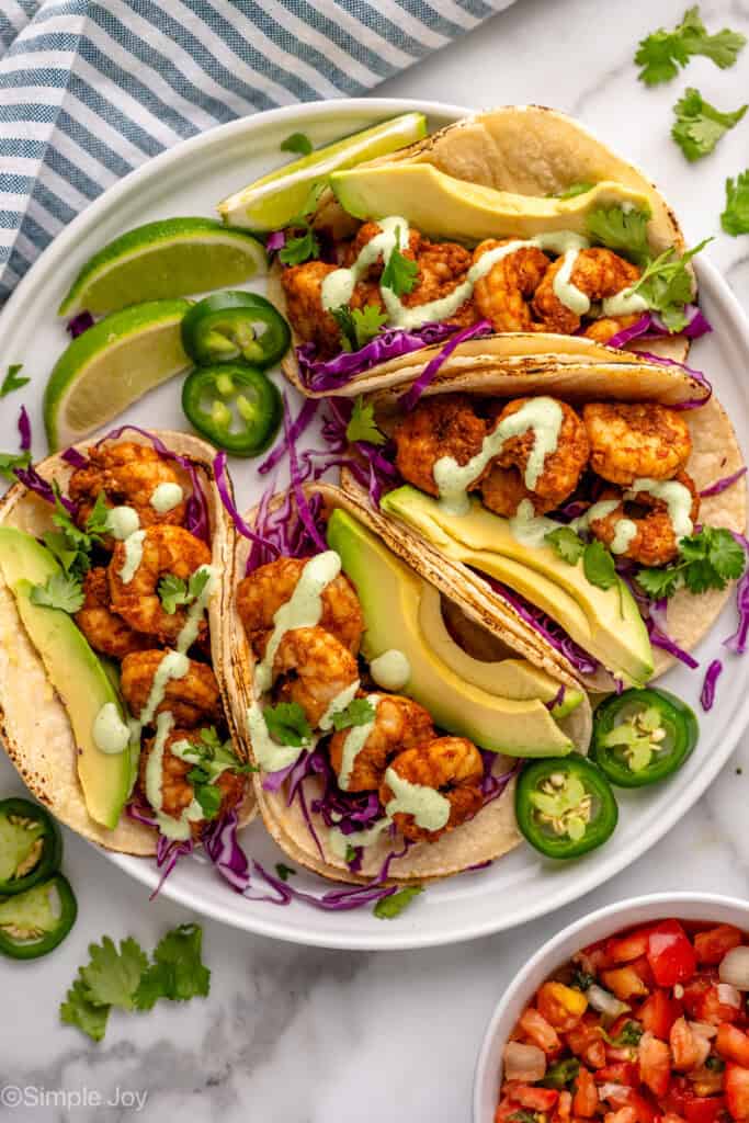 overhead of a plate of four shrimp tacos with avocado, fresh cilantro, red cabbage, and cilantro lime crema sauce