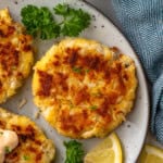 overhead of plate of crab cakes with fresh parsley and lemon slices. Cup of sauce sitting beside.