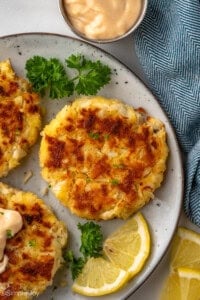 overhead of plate of crab cakes with fresh parsley and lemon slices. Cup of sauce sitting beside.