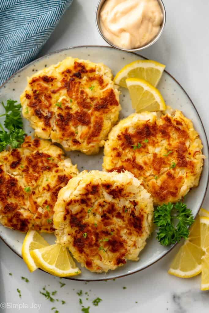 overhead of plate of crab cakes with fresh parsley and lemon slices. Cup of sauce sitting beside.
