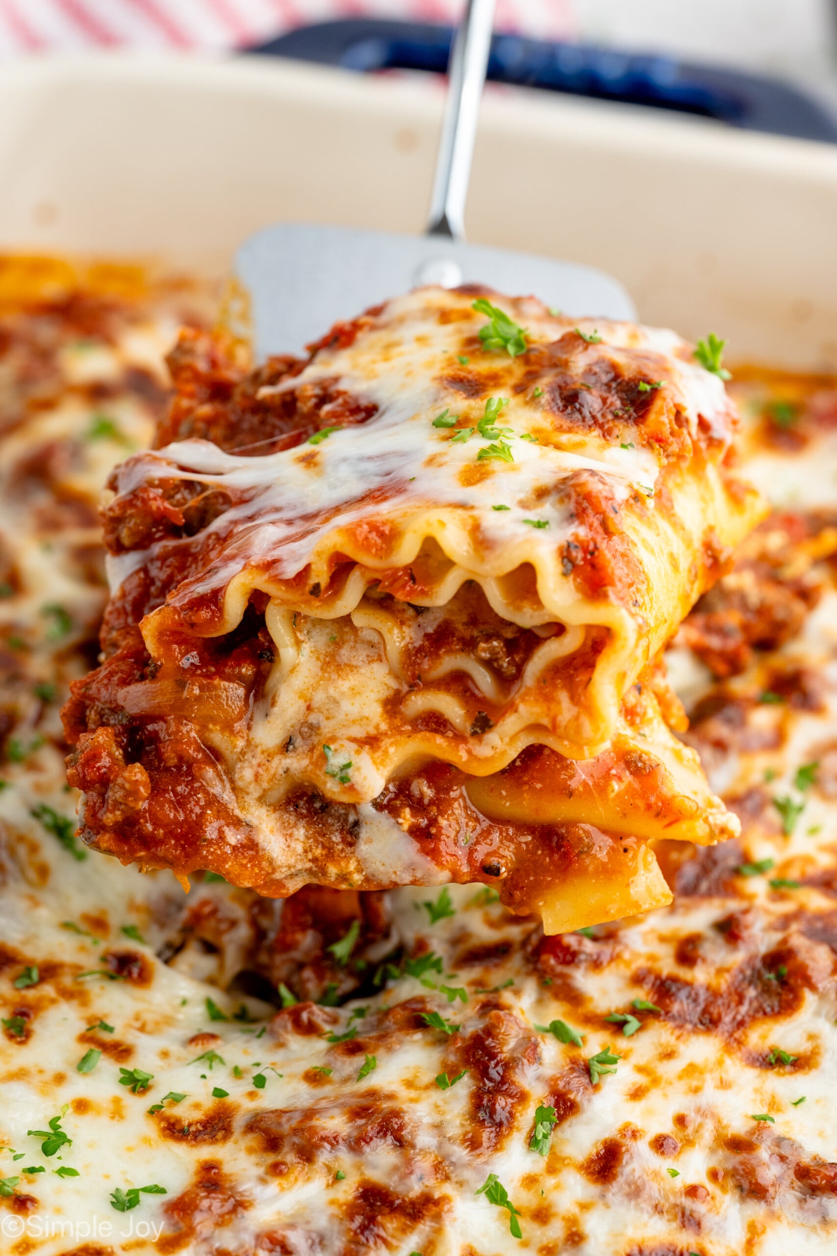 Serving spatula lifting Lasagna Roll Up from baking dish of Lasagna Roll Ups recipe, garnished with fresh parsley