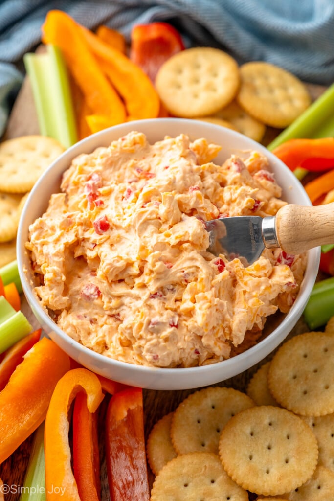 Bowl of Pimento Cheese with spreading knife, surrounded by platter of crackers and sliced vegetables for dipping.
