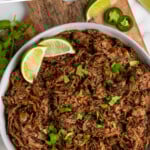 Pinterest graphic for beef barbacoa. Image shows overhead of white bull of beef barbacoa garnished with lime wedges and cilantro. Says "slow cooker beef barbacoa simplejoy.com"