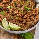 Pinterest graphic for beef barbacoa. Image shows white bull full of beef barbacoa, garnished with cilantro and lime wedges with two forks. Says "slow cooker beef barbacoa simplejoy.com"