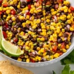 Pinterest graphic of Black Bean and Corn Salsa. Image shows a bowl of Black Bean and Corn Salsa with lime wedges. Tortilla chips, fresh cilantro, and lime wedges surrounding. Says "super easy Black Bean and Corn Salsa simplejoy.com"