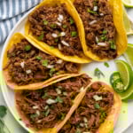 Overhead of beef barbacoa in corn tortillas garnished with diced onion and cilantro, lime wedges, cilantro, and diced jalapeños sitting beside