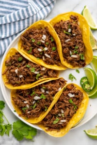 Overhead of beef barbacoa in corn tortillas garnished with diced onion and cilantro, lime wedges, cilantro, and diced jalapeños sitting beside