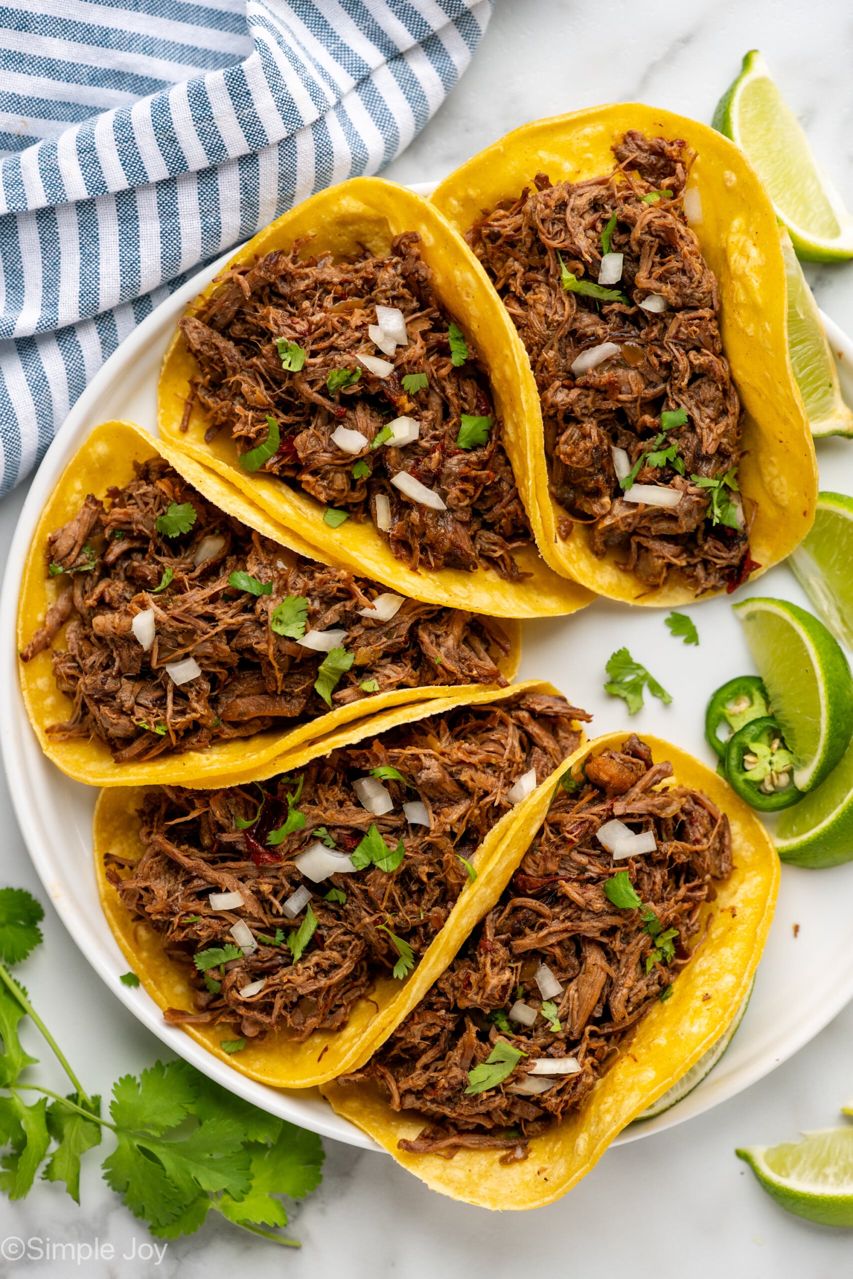 Overhead of beef barbacoa in corn tortillas garnished with diced onion and cilantro, lime wedges, cilantro, and diced jalapeños sitting beside