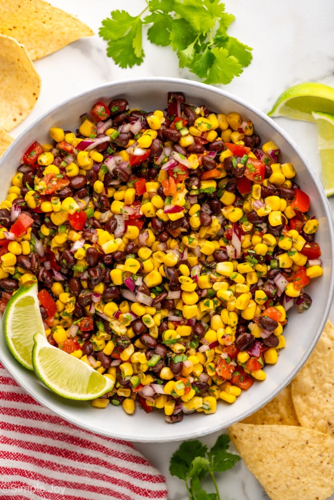 Overhead of bowl of Black Bean and Corn Salsa with lime wedges, fresh cilantro, and tortilla chips surrounding