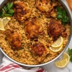 Overhead of skillet Chicken and Rice, garnished with lemon slices and parsley. Lemons slices, parsley, and bread sitting beside.