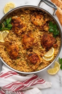 Overhead of skillet Chicken and Rice, garnished with lemon slices and parsley. Lemons slices, parsley, and bread sitting beside.